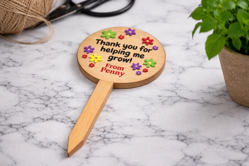 May include: Wooden garden stake with a round top and a pointed base. The top is decorated with colorful flower accents and the text "Thank you for helping me grow! From Penny". A plant in a pot is in the background.