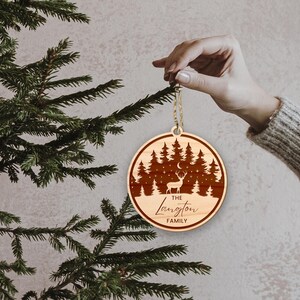 Op de afbeelding: Een ronde houten kerstversiering met een lasergesneden ontwerp van een hert dat in een bos staat met een halve maan en sterren. De versiering is gepersonaliseerd met de tekst "THE Langton FAMILY".