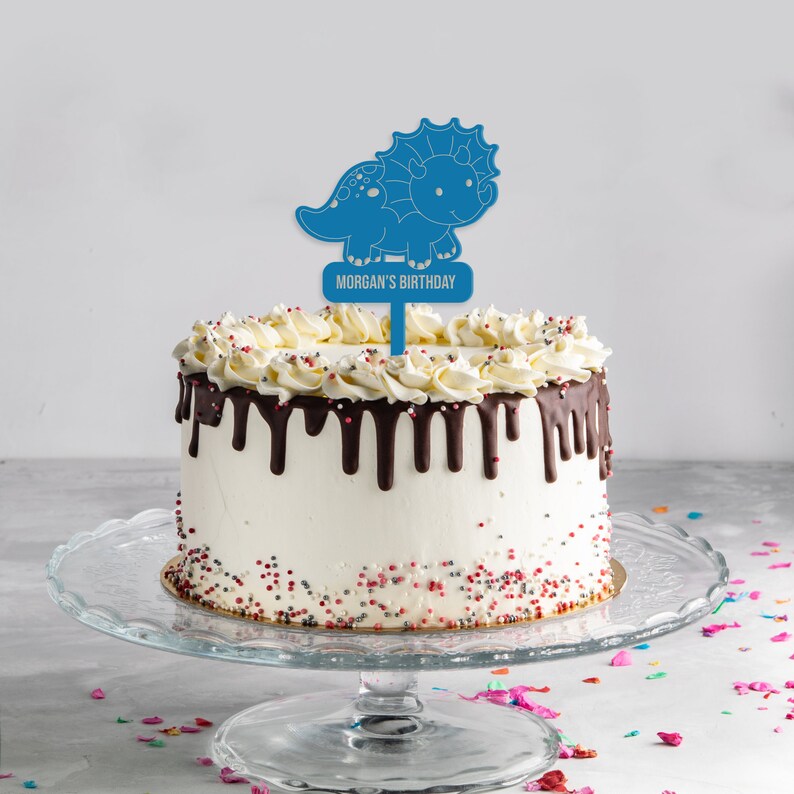 May include: A white cake with chocolate frosting and sprinkles topped with a blue dinosaur cake topper that says "Morgan's Birthday".