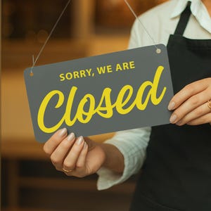 Op de afbeelding: Een rechthoekig grijs bord met de woorden "SORRY, WE ARE" in het wit en "Closed" in gele letters. Het bord wordt vastgehouden door een persoon die een zwarte schort draagt.