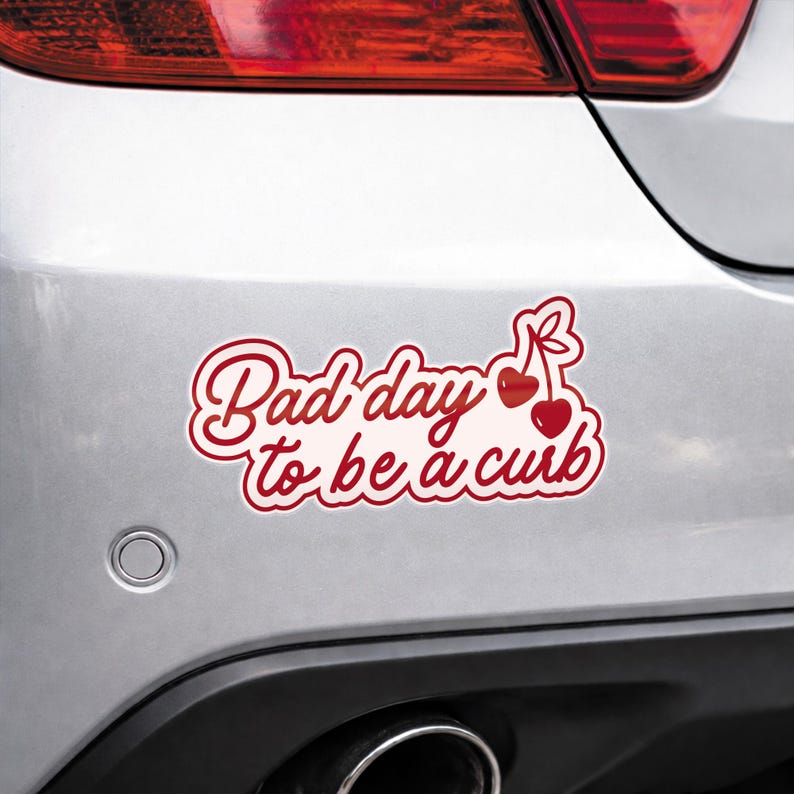 K&ouml;nnte beinhalten: Roter und wei&szlig;er Aufkleber mit dem Schriftzug "Bad day to be a curb" in Schreibschrift. Zwei rote Kirschen mit gr&uuml;nen Stielen befinden sich auf der rechten Seite. Der Aufkleber befindet sich auf der R&uuml;ckseite eines silbernen Autos.