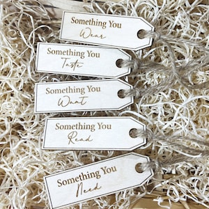 Pu&ograve; includere: Cinque etichette regalo in legno con un cordoncino attaccato. Ogni etichetta ha il testo "Something You" seguito da una parola diversa: "Wear", "Taste", "Want", "Read" e "Need".