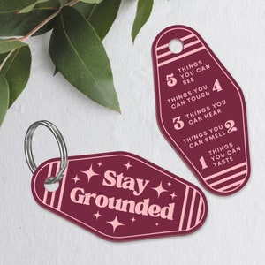 May include: A maroon keychain with a pink and white striped design. The keychain has a metal ring and a tag that says "Stay Grounded". The other tag has a list of five things you can experience with your senses: see, touch, hear, smell, and taste.