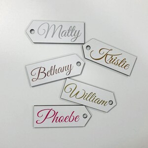 May include: Five white wooden gift tags with different names written in gold, silver, pink, and rose gold lettering. The names are Matty, Kristie, Bethany, William, and Phoebe.