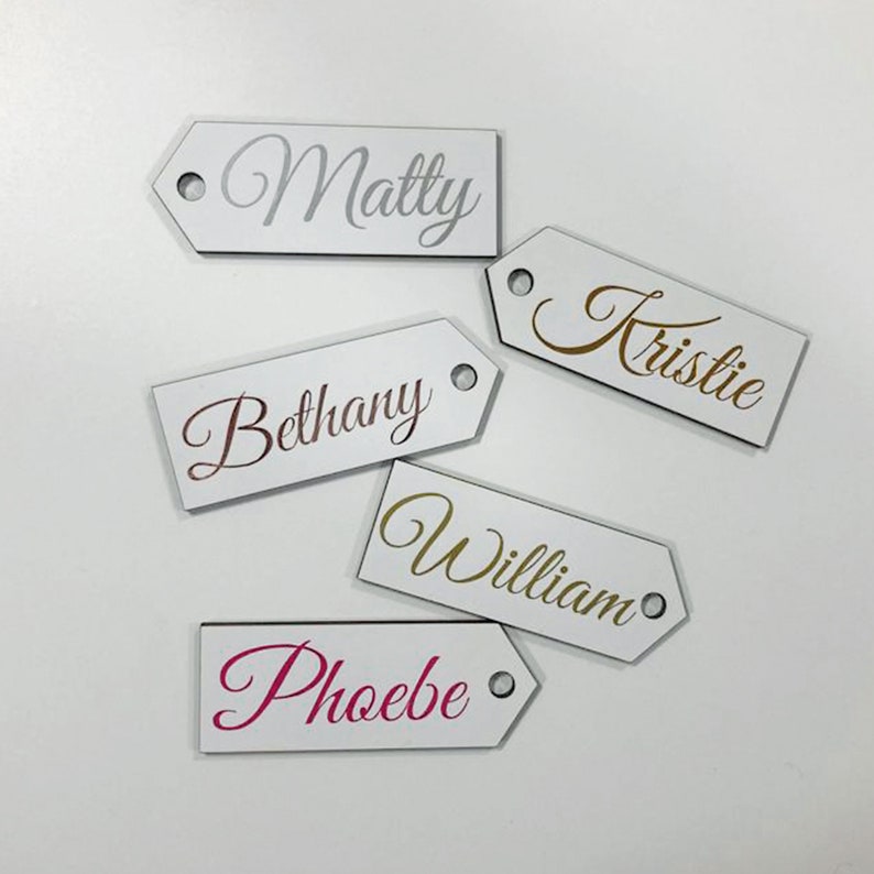May include: Five white wooden gift tags with different names written in gold, silver, pink, and rose gold lettering. The names are Matty, Kristie, Bethany, William, and Phoebe.