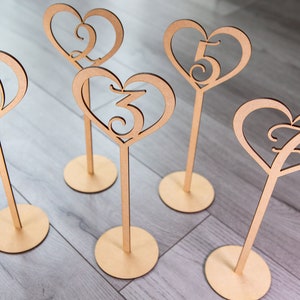 May include: Set of five wooden heart-shaped table numbers, each with a different number from 1 to 5, on a wooden stand. The numbers are cut out of the hearts and are in a cursive font.