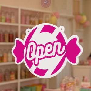 May include: A white and pink candy-shaped sign with the word "Open" in a cursive font. The sign is hanging from a clear suction cup. The background shows a candy store with shelves of colorful treats.
