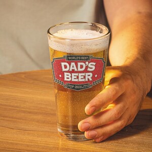 May include: A clear pint glass filled with beer and foam. The glass has a red and white label that reads "World's Best Dad's Beer Top Quality". The glass is held by a person.