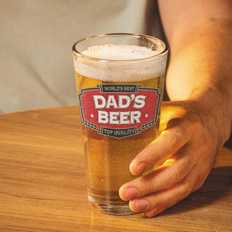 May include: A clear pint glass filled with beer and foam. The glass has a red and white label that reads "World's Best Dad's Beer Top Quality". The glass is held by a person.