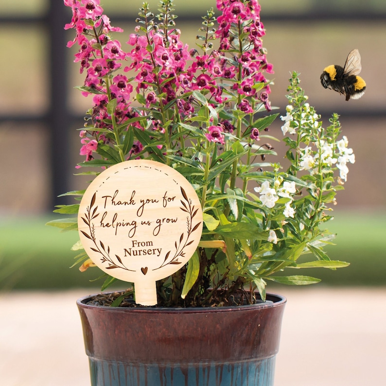 May include: Wooden plant stake with a thank you message for helping us grow from nursery. The stake is in a pot with pink and white flowers. A bumblebee is flying near the flowers.