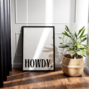 Op de afbeelding: Zwart-witte print met het woord "HOWDY" in een vet lettertype, ingelijst in een zwart frame, hangend aan een witte muur met een plant in een geweven mand in de voorgrond.