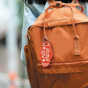 May include: A red keychain with a white cross and the text "Medical Info!" hanging from a brown backpack.