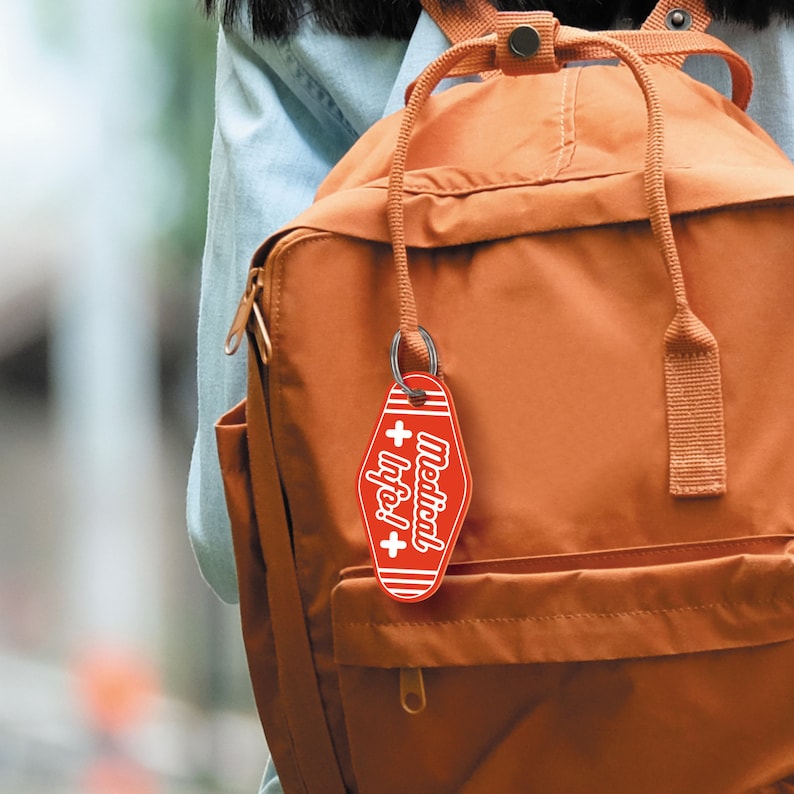 May include: A red keychain with a white cross and the text "Medical Info!" hanging from a brown backpack.