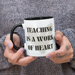 K&ouml;nnte beinhalten: Eine wei&szlig;e Keramiktasse mit schwarzem Henkel und schwarzem Rand. Die Tasse hat ein graues Herz mit dem Text "TEACHING IS A WORK OF HEART" in Schwarz.