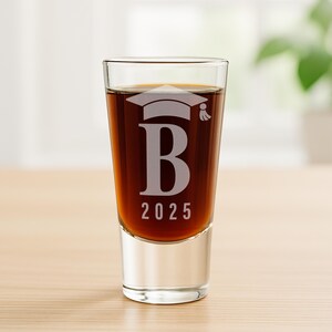 K&ouml;nnte beinhalten: Ein klares Schnapsglas, gef&uuml;llt mit einer dunklen Fl&uuml;ssigkeit. Das Glas ist mit einer Abschlusskappe, dem Buchstaben "B" und dem Jahr "2025" graviert. Das Glas steht auf einer hellbraunen Holzoberfl&auml;che.