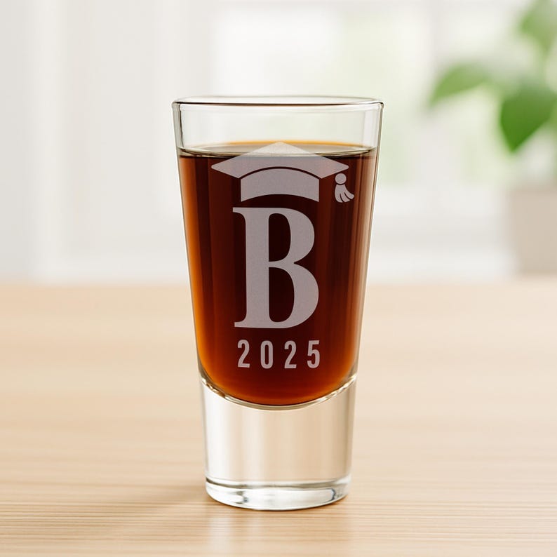 K&ouml;nnte beinhalten: Ein klares Schnapsglas, gef&uuml;llt mit einer dunklen Fl&uuml;ssigkeit. Das Glas ist mit einer Abschlusskappe, dem Buchstaben "B" und dem Jahr "2025" graviert. Das Glas steht auf einer hellbraunen Holzoberfl&auml;che.
