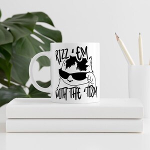May include: White ceramic mug featuring a black and white illustration of a cat wearing sunglasses and the text "Rizz 'em with the -tism".