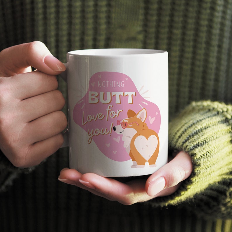 May include: White ceramic mug featuring a cartoon corgi with heart-shaped markings and the text "Nothing Butt Love for you!" in pink and gold. The corgi is wearing glasses. The mug is held by a person.