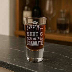 May include: A clear shot glass filled with a brown liquid, featuring the etched text "YOU GAVE IT YOUR BEST SHOT & NOW YOU'RE A GRADUATE". The glass is on a dark countertop, with a blurred background of bottles and glassware.