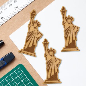 May include: Three wooden cutout decorations of the Statue of Liberty. The decorations are brown and have a rustic, distressed look.