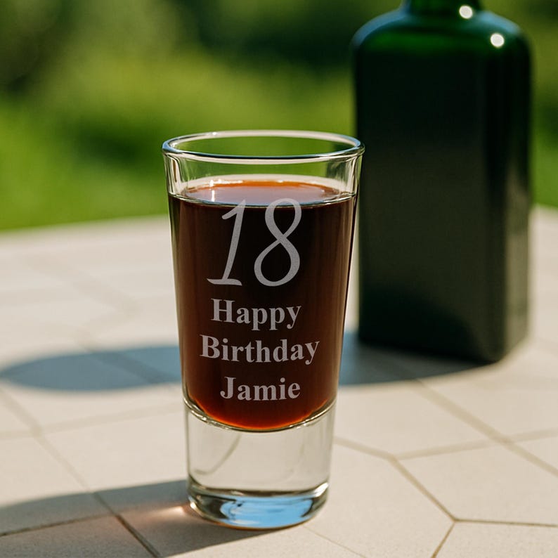 May include: A clear shot glass filled with a dark liquid, featuring the etched text "18 Happy Birthday Jamie." A green bottle is blurred in the background. The glass is on a light-colored surface, with a green background.
