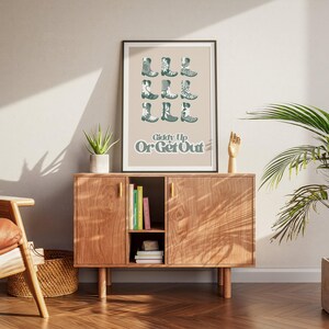 May include: A beige poster with a graphic of cowboy boots in a green color. The text on the poster reads "Giddy Up Or Get Out". The poster is hanging on a wall above a wooden cabinet.