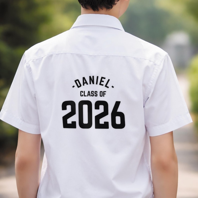 May include: White short-sleeved shirt with the name "DANIEL" and "CLASS OF 2026" printed in black on the back. The shirt has a classic collar and a button-down front. The background is blurred, suggesting an outdoor setting.