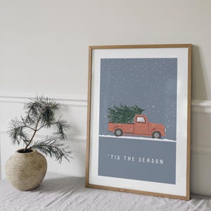 May include: A framed print of a red pickup truck carrying a Christmas tree against a blue snowy background. The text 'Tis the Season is printed below the truck.