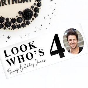 May include: A white banner with the text "LOOK WHO'S 40 Happy Birthday James" and a photo of a smiling man. The number 40 is in large black font. The banner is decorated with black confetti and a decorated cake.