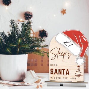 May include: A wooden countdown sign with a Santa hat design. The sign says "Sleep's Until Santa Visits Our Home" and has a space to write in the number of sleeps until Christmas. The sign is standing on a table with a black marker next to it.