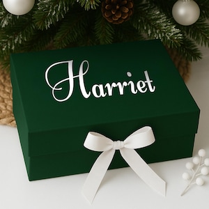 K&ouml;nnte beinhalten: Eine dunkelgr&uuml;ne Geschenkbox mit dem Namen "Harriet" in wei&szlig;er Schrift auf dem Deckel. Ein wei&szlig;es Band ist zu einer Schleife um die Box gebunden. Die Box ist mit Weihnachtsschmuck und Gr&uuml;n geschm&uuml;ckt.