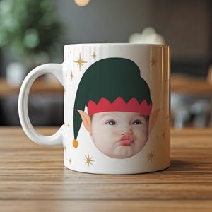 May include: White ceramic mug featuring a baby's face wearing an elf hat with a green cap and red trim. The mug has a white handle and is decorated with gold stars. A festive holiday-themed gift.