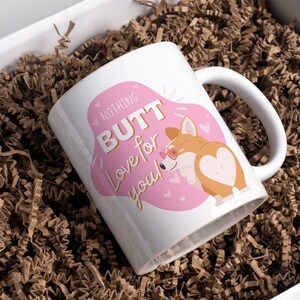 May include: White ceramic mug with a handle, featuring a pink and white design with the text "NOTHING BUTT Love for you!". The design includes a cartoon corgi wearing sunglasses. The mug is presented in a white gift box with brown paper filler.