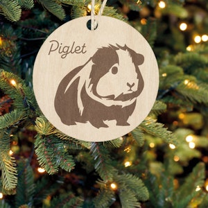 May include: A round wooden ornament with a brown and white guinea pig silhouette and the word "Piglet" written above it.