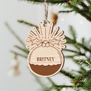 May include: A wooden Christmas ornament shaped like a pudding with a holly sprig and berries. The ornament is personalized with the name "BRITNEY".