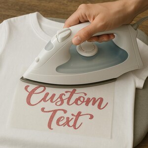 May include: A white iron is being used to press a pink glitter iron-on design onto a white t-shirt. The design says "Custom Text".