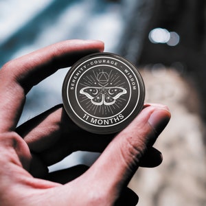 May include: A black round coin with white text and a moth design. The coin reads "SERENITY - COURAGE - WISDOM" around the edge and "11 MONTHS" at the bottom. A moth with moon and star details is in the center.
