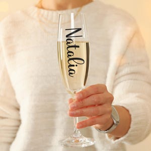 May include: A clear glass champagne flute with a black vinyl sticker that says "Natalia" on it. The flute is filled with champagne and is being held by a person.