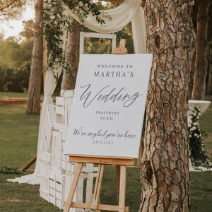 K&ouml;nnte beinhalten: Ein wei&szlig;es Hochzeitsschild mit schwarzem Text, der lautet "Willkommen zur Hochzeit von Martha mit Josh Wir freuen uns, dass Sie da sind. 28/11/23". Das Schild steht auf einem Staffelei vor einem Baum und wei&szlig;en Holzkisten.