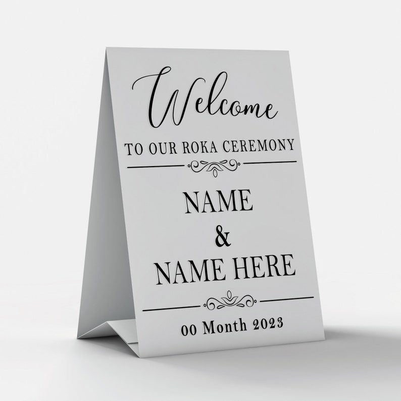May include: A white table sign with black text that reads "Welcome to our Roka Ceremony NAME & NAME HERE 00 Month 2023".