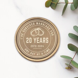 Puede incluir: Placa redonda de madera con la inscripci&oacute;n "I've survived marriage for... 20 years ESTD 2004 One day at a time." El dise&ntilde;o incluye dos anillos entrelazados. La placa es de color marr&oacute;n claro. Hojas de eucalipto en el fondo.