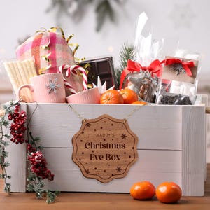 May include: A white wooden crate filled with a Christmas Eve box gift set. The crate has a wooden sign hanging from it that says "Maddy's Christmas Eve Box". The box contains a variety of items, including a pink mug, a blanket, candy canes, chocolate, and oranges.