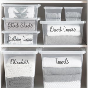 May include: Clear plastic storage bins labeled with black text: "Fitted Sheets", "Pillow Cases", "Duvet Covers", "Blankets", and "Towels". The bins are stacked on white shelves and contain folded linens.