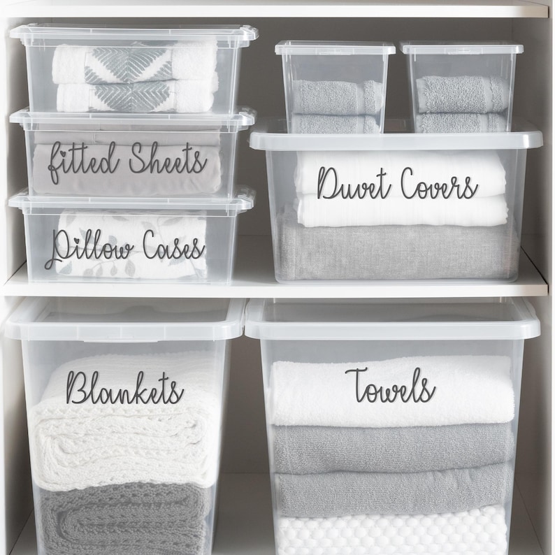 May include: Clear plastic storage bins labeled with black text: "Fitted Sheets", "Pillow Cases", "Duvet Covers", "Blankets", and "Towels". The bins are stacked on white shelves and contain folded linens.