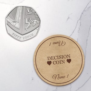 May include: A silver fifty pence coin and a round wooden decision coin. The fifty pence coin features a detailed design. The wooden coin has the words "DECISION COIN" and "Name 1" engraved on it, with two heart symbols.