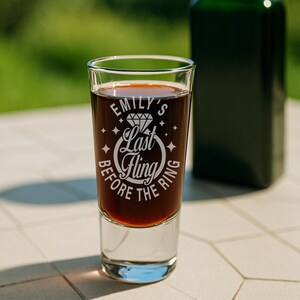 May include: A clear shot glass filled with a dark liquid, with the text "Emily's Last Fling Before the Ring" in white. The glass is on a white surface, with a blurred green background. A dark green bottle is in the background.