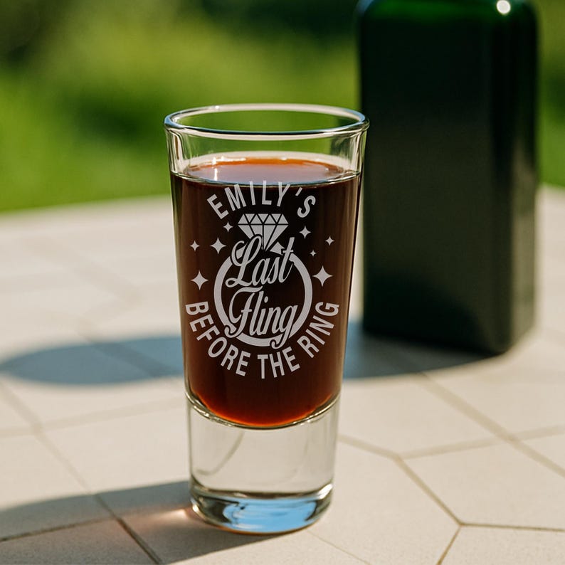 May include: A clear shot glass filled with a dark liquid, with the text "Emily's Last Fling Before the Ring" in white. The glass is on a white surface, with a blurred green background. A dark green bottle is in the background.
