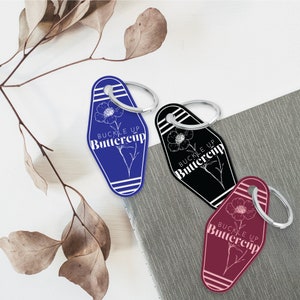 May include: Three keychains with a motel key design. Each keychain features a white outline of a flower with the text "Buckle Up Buttercup" in white. The keychains are blue, black, and burgundy.