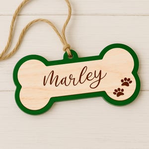 Może przedstawiać: Drewniana zawieszka w kształcie kości dla psa z imieniem "Marley" w brązowym skrypcie. Zawieszka ma zieloną obw&oacute;dkę i dwa brązowe odciski łap. Do zawieszenia przymocowana jest pętla z naturalnego sznurka.