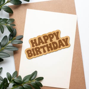 May include: A white card with a brown "Happy Birthday" sign on it. The card is surrounded by green leaves.
